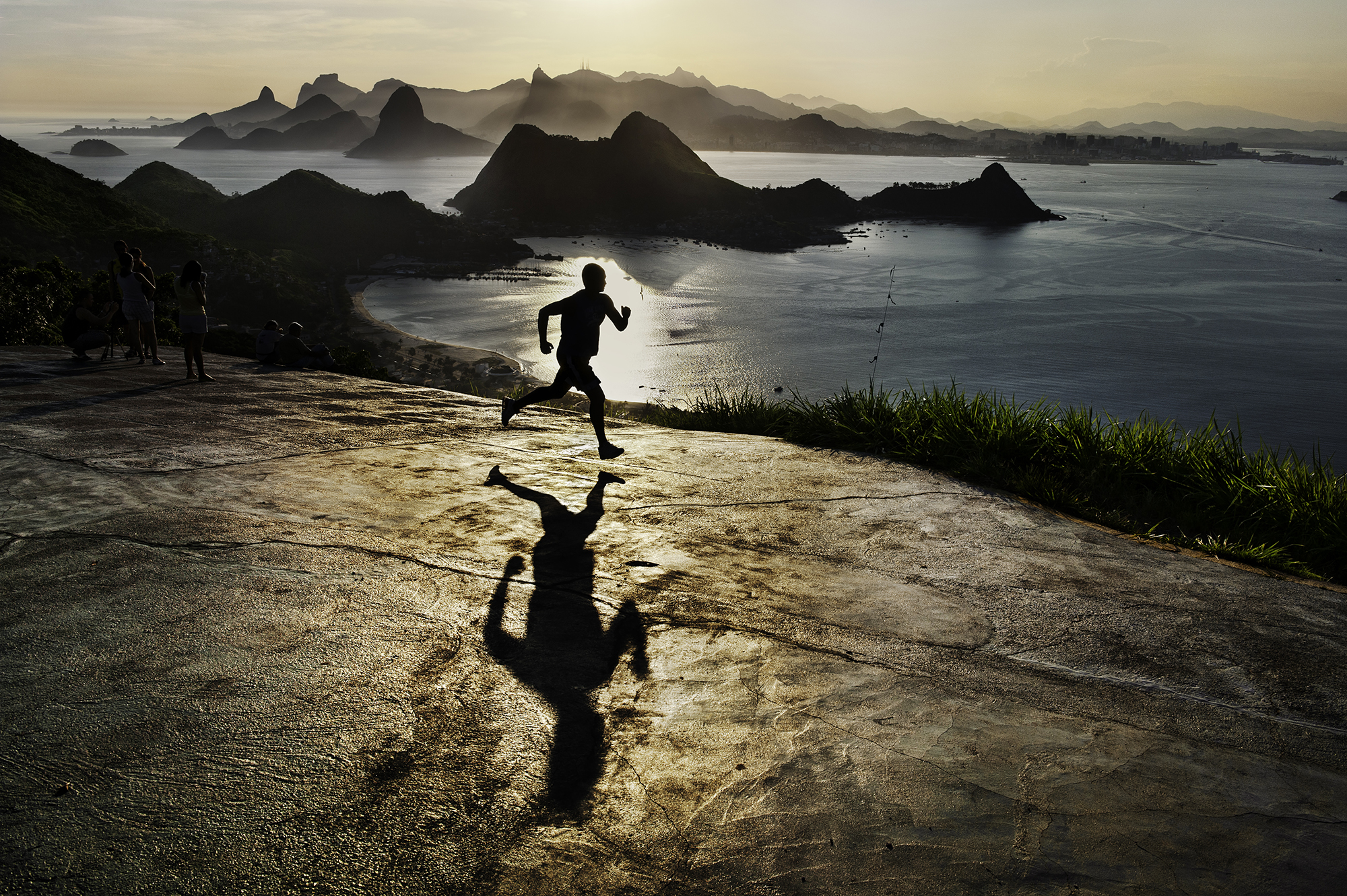 In Steve McCurry's 2009 photograph of a man running at dawn in Rio de Janeiro, Brazil, the early morning atmosphere is captured with McCurry’s trademark sense of color and composition. The runner, silhouetted against the warm, golden hues of the sunrise, moves along a coastline that is both vast and serene, with Rio’s iconic mountains rising majestically in the background. The image conveys a powerful sense of solitude and focus, as the lone figure embraces the peaceful quiet of dawn in one of the world’s most vibrant cities. McCurry’s eye for detail highlights not only the natural beauty of the scene but also the universal human drive for perseverance and inner strength. This image, like many of McCurry’s works, merges the beauty of the landscape with a moment of human resilience, creating a scene that feels both intimate and expansive.