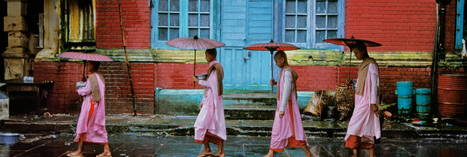 Procession of Nuns, Rangoon, Burma, 1994, presents an image inundated with vibrant colours of orange, pink, yellow and blue to contrast a serene and still moment. There is also a juxtaposition of colour with the darker pavement, allowing a contrast that further enhances the image’s potency. Framing the four figures, the composition is balanced. The nuns hold rice bowls for the locals to offer them food with which they will return to the nunnery. The blue door in the background acts as a focal point, drawing the viewers gaze to the centre of the image. Our eye is then led around the work by the linear formation created by the procession and their opened umbrellas, encouraging us to survey every aspect of the work.