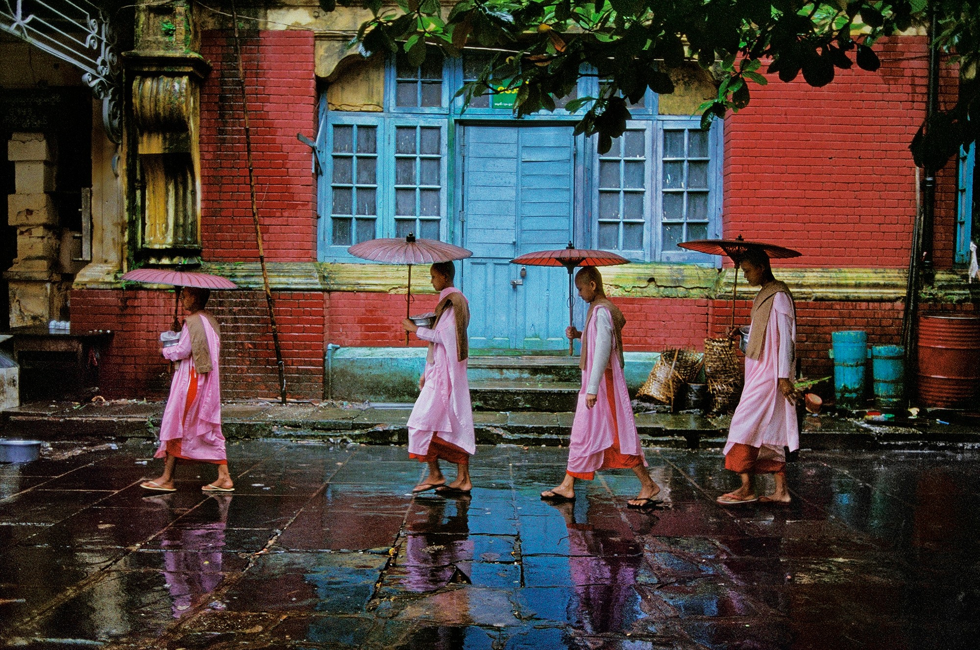 Procession of Nuns, Rangoon, Burma, 1994, presents an image inundated with vibrant colours of orange, pink, yellow and blue to contrast a serene and still moment. There is also a juxtaposition of colour with the darker pavement, allowing a contrast that further enhances the image’s potency. Framing the four figures, the composition is balanced. The nuns hold rice bowls for the locals to offer them food with which they will return to the nunnery. The blue door in the background acts as a focal point, drawing the viewers gaze to the centre of the image. Our eye is then led around the work by the linear formation created by the procession and their opened umbrellas, encouraging us to survey every aspect of the work.